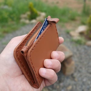 May include: A brown leather wallet with a zipper closure and a card peeking out. The wallet is open, revealing the interior and a card with the text "CLUB". The stitching is visible, and the leather has a smooth texture.