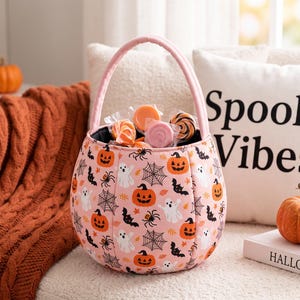 May include: A round, soft-sided fabric bucket with a pink handle and a light pink exterior featuring a repeating pattern of orange jack-o'-lanterns, white ghosts, black bats, black spiders, and spiderwebs, filled with several individually wrapped sweets.