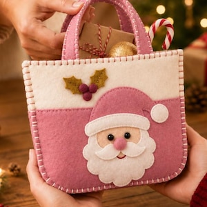 Pode incluir: Uma bolsa de feltro rosa e creme com uma aplicação do rosto do Pai Natal na frente, decorada com folhas de azevinho e bagas. A bolsa tem costuras brancas visíveis ao longo das bordas e contém um pequeno presente e um bastão doce.