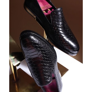 May include: A pair of black slip-on loafers featuring a textured snakeskin pattern. The shoes have a glossy finish, a rounded toe, and a metallic accent on the heel. The interior of the shoes is lined with a bright pink material.