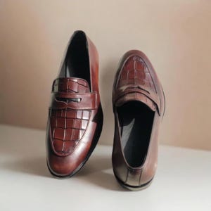 Pode incluir: Um par de mocassins de couro castanho. Os sapatos apresentam um design clássico de mocassim com um padrão texturizado, semelhante ao de um crocodilo, na gáspea. O couro tem um acabamento polido e os sapatos estão sobre um fundo neutro.