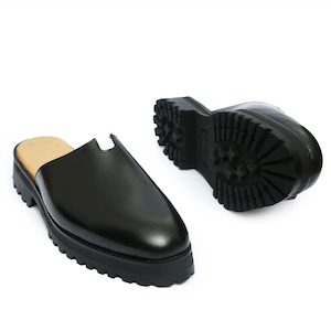 Könnte beinhalten: Ein Paar schwarze Slipper-Mules mit glattem, poliertem Obermaterial und einem gekerbten Kragen. Das Schuhwerk verfügt über eine dicke, klobige Plateausohle mit einem tiefen, robusten Profil und einem beigefarbenen Innenfutter.