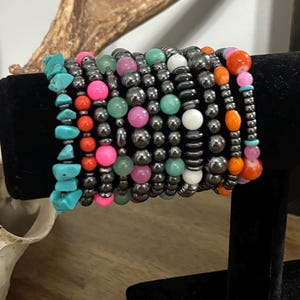 May include: A collection of stacked beaded bracelets featuring a variety of materials including turquoise-colored stone chips, metallic dark gray spheres, and smooth round beads in vibrant shades of neon pink, orange, green, white, and light pink.