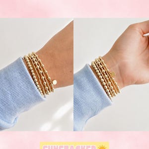 Könnte beinhalten: Ein Set aus fünf dehnbaren Armbändern mit goldfarbenen Perlen am Handgelenk. Jedes Armband besteht aus kleinen, gleichmäßigen Metallkugeln, wobei an einem Armband ein kleiner, flacher, kreisförmiger goldfarbener Anhänger befestigt ist.