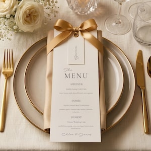 May include: A white menu card with black text titled The Menu, featuring sections for Appetizer, Entree, and Dessert. The card is adorned with a gold satin ribbon bow and a small name tag, resting on a beige napkin atop a cream-colored plate.