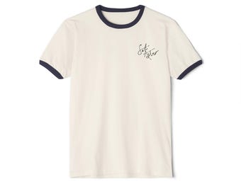May include: Cream-colored ringer t-shirt with navy blue trim around the collar and sleeves. The shirt features the handwritten text "Set Astro" in black on the front. A casual, everyday wear tee.