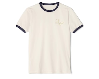 May include: Cream-colored ringer t-shirt with navy blue trim on the collar and sleeves. The shirt features gold script text that reads "Soft Silver" on the upper left chest. The t-shirt is made of a soft material.