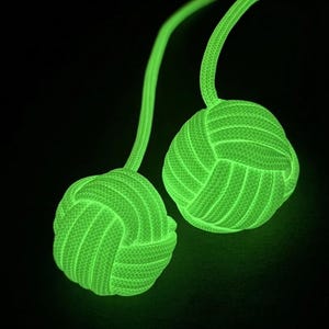 May include: Two spherical monkey fist knots crafted from bright neon green paracord. Each knot is attached to a matching green cord, creating a pair of luminous, textured spheres that glow against a dark background, showcasing intricate woven patterns.