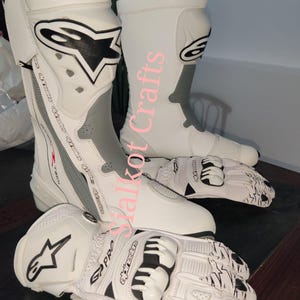 Könnte beinhalten: Weiß-graues Motorradstiefel- und Handschuh-Set. Die hohen Stiefel haben ein schwarzes Logo und graue Akzente. Die weißen Handschuhe haben schwarze Details und ein passendes Logo. Entwickelt für Schutz und Leistung beim Motorradfahren.