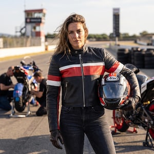 May include: A black leather motorcycle jacket featuring a horizontal red and white stripe across the chest and sleeves, a full-length front zipper, and quilted shoulder detailing, paired with dark grey denim jeans and black leather gloves.