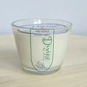 May include: A clear glass jar candle filled with off-white wax. A label on the front features the text "HAND POURED COCOSOY CANDLE" and "Divine by Jilly" in green, accompanied by a small botanical illustration of lavender sprigs.