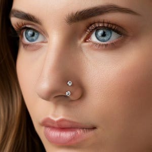 May include: A close-up shot of a person's face, highlighting two small, sparkling nose studs. The studs are set in a silver or white metal. The person has fair skin, blue eyes, and brown eyebrows. The focus is on the nose and the jewellery.
