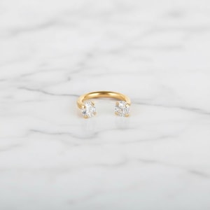 May include: A gold-colored ring with two clear, round-cut gemstones. The ring has a unique open design, with the gemstones set at opposite ends of the band. The ring is displayed on a white marble surface.