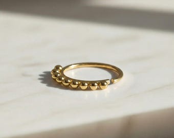 May include: A gold-colored ring with a row of small, round beads along the top. The ring is set against a white marble surface, with soft shadows adding depth. The jewelry piece is simple and elegant.