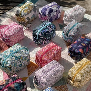 May include: A selection of patterned cosmetic bags. Each bag has a rectangular shape with a zip closure and a small handle. The bags showcase floral and abstract designs in shades of pink, blue, purple, and yellow. The bags are arranged on a patterned surface.