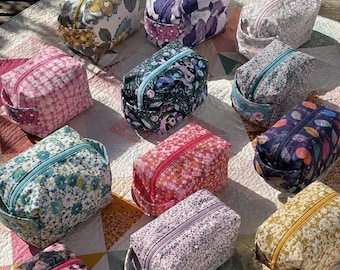 May include: A collection of colorful, patterned cosmetic bags. Each bag features a rectangular shape with a zippered closure and a small handle. The bags display various floral and abstract designs in shades of pink, blue, purple, and yellow.
