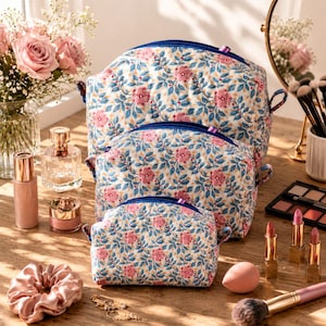 May include: A set of three quilted cosmetic bags in varying sizes, featuring a repeating pattern of pink flowers and blue leaves on a white background. Each bag has a blue zip closure and small side tabs, arranged in a stacked formation.