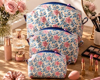 May include: A set of three quilted cosmetic bags in varying sizes, featuring a repeating pattern of pink flowers and blue leaves on a white background. Each bag has a blue zipper closure and small side tabs, arranged in a stacked formation.