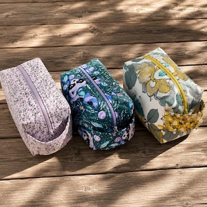 May include: Three rectangular cosmetic bags with zipper closures. The bags feature floral patterns in lavender, teal, and yellow. Each bag has a small handle on one side. The bags are arranged on a wooden surface.