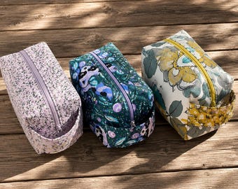 May include: Three rectangular cosmetic bags with zipper closures. The bags feature floral patterns in lavender, teal, and yellow. Each bag has a small handle on one side. The bags are arranged on a wooden surface.