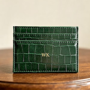 May include: A dark green card holder with a crocodile skin texture. The card holder has multiple slots and the initials "WK" in gold lettering. The card holder is rectangular and has stitched edges.