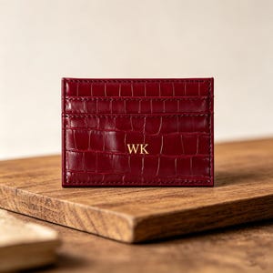 Puede incluir: Un tarjetero burdeos con textura de cocodrilo. El tarjetero tiene múltiples ranuras y las iniciales "WK" en letras doradas. El tarjetero es rectangular y está sobre una superficie de madera.