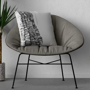 May include: A square throw pillow with a light gray background featuring a black and white graphic illustration of a stack of books on the left side and the vertical text "TSUNDOKU" printed in black on the right side of the pillow cover.