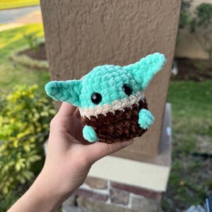 May include: A handmade crochet plush toy in the likeness of a character with large ears. The toy features a teal head and ears, a brown body, and black eyes. The plush is held in a person's hand.