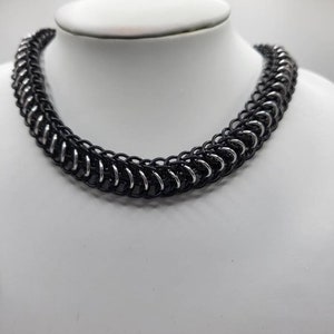 May include: A black and silver chainmail necklace. The chainmail is made of small metal rings that are interlocked to create a strong and flexible material.