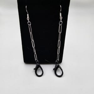 May include: A pair of silver-colored chain mask holders with black lobster clasps. The chains are made of elongated links and are suspended from silver-colored ear hooks. The mask holders are displayed against a black background.