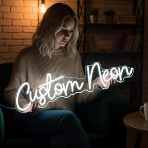 Pu&ograve; includere: Un'insegna al neon bianca con le parole "Custom Neon" in un carattere corsivo. L'insegna &egrave; tenuta da una persona in una stanza con un muro di mattoni e un'illuminazione calda.