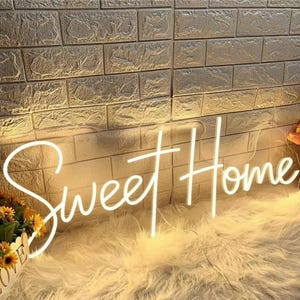 Pu&ograve; includere: Un'insegna al neon bianca con la scritta "Sweet Home" in corsivo. &Egrave; montata su un muro di mattoni strutturato. L'insegna &egrave; posta su una superficie bianca e soffice, con fiori in un cestino.