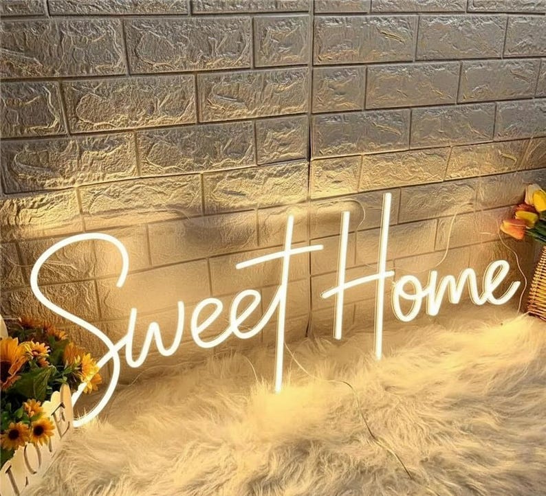 Pu&ograve; includere: Un'insegna al neon bianca con la scritta "Sweet Home" in corsivo. &Egrave; montata su un muro di mattoni strutturato. L'insegna &egrave; posta su una superficie bianca e soffice, con fiori in un cestino.