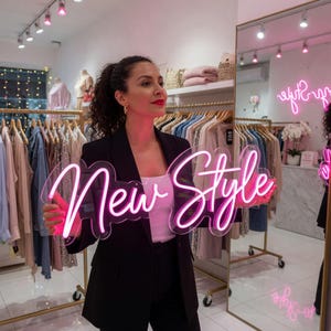 Pu&ograve; includere: Una donna tiene in mano un'insegna al neon rosa con la scritta "New Style". L'insegna ha un supporto trasparente. Sullo sfondo si vedono appendiabiti e uno specchio, che indicano un ambiente di boutique di moda.