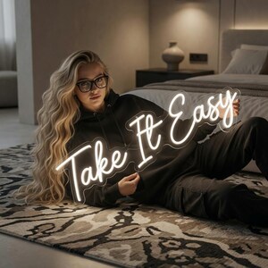 May include: A white neon sign featuring the cursive text Take It Easy, constructed with flexible LED tubing mounted on a clear acrylic backing. The sign emits a bright, steady glow, displaying the words in a flowing, connected script style.
