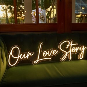Pu&ograve; includere: Un'insegna al neon bianca calda con la scritta "Our Love Story" in corsivo. L'insegna &egrave; posizionata su un divano di velluto verde scuro, con una finestra e uno scenario esterno sullo sfondo. Un elemento decorativo per matrimoni o eventi.