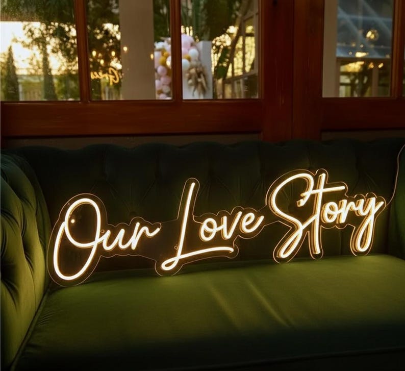 Pu&ograve; includere: Un'insegna al neon bianca calda con la scritta "Our Love Story" in corsivo. L'insegna &egrave; posizionata su un divano di velluto verde scuro, con una finestra e uno scenario esterno sullo sfondo. Un elemento decorativo per matrimoni o eventi.