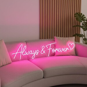 Pu&ograve; includere: Un'insegna al neon rosa con la scritta "Always & Forever" in corsivo, con un simbolo a forma di cuore. L'insegna &egrave; posizionata su un divano grigio con cuscini rosa. Sullo sfondo una parete a doghe di legno e una pianta.