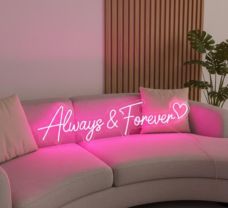 Pu&ograve; includere: Un'insegna al neon rosa con la scritta "Always & Forever" in corsivo, con un simbolo a forma di cuore. L'insegna &egrave; posizionata su un divano grigio con cuscini rosa. Sullo sfondo una parete a doghe di legno e una pianta.