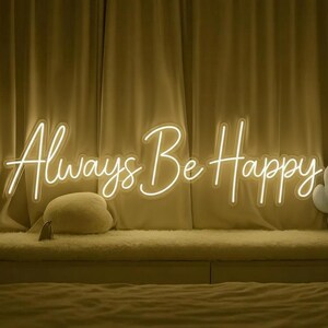 Pu&ograve; includere: Un'insegna al neon con la scritta "Always Be Happy" in corsivo. L'insegna emette una luce bianca calda ed &egrave; montata su uno sfondo di tende beige. Un cuscino soffice e una piccola etichetta sono visibili sotto l'insegna.