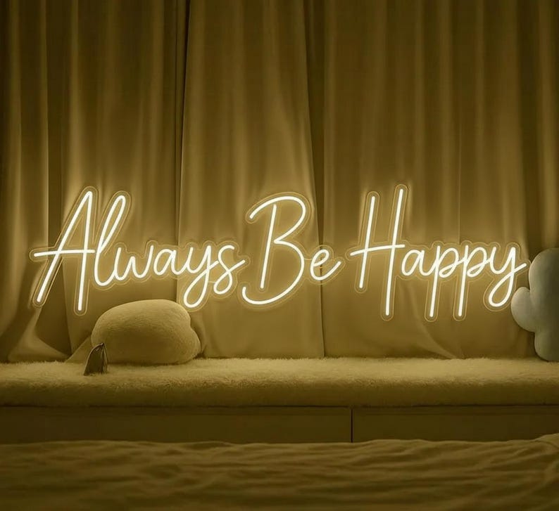 Pu&ograve; includere: Un'insegna al neon con la scritta "Always Be Happy" in corsivo. L'insegna emette una luce bianca calda ed &egrave; montata su uno sfondo di tende beige. Un cuscino soffice e una piccola etichetta sono visibili sotto l'insegna.