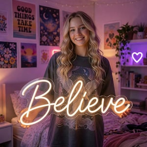 May include: A clear acrylic neon sign featuring the word Believe in a glowing warm white cursive script. The sign is shaped to follow the contour of the lettering and is held by a person wearing a dark gray t-shirt with a graphic print.