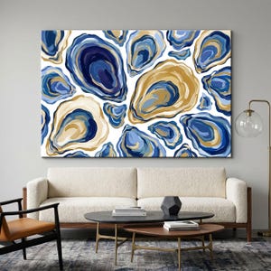 Op de afbeelding: Een abstracte canvas kunstprint met een patroon van oesterschelpvormen in tinten blauw en goud tegen een witte achtergrond. Het kunstwerk wordt weergegeven boven een crèmekleurige bank in een woonkamer.
