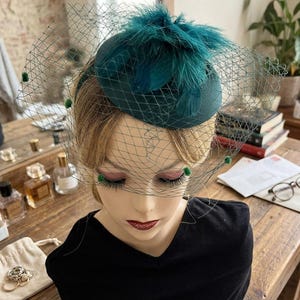 May include: A dark teal fascinator hat featuring a rounded base, a cluster of matching feathers on top, and a wide-reaching green birdcage veil accented with small green pom-poms, displayed on a mannequin head.