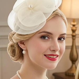 May include: A cream-colored fascinator hat featuring layered sheer fabric petals and a central pearl accent. The headpiece is worn with a matching pearl necklace and pearl drop earrings, creating a coordinated set of accessories with a smooth finish.
