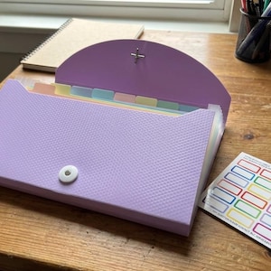 May include: A lavender expanding file folder with a textured surface, open to reveal multi-colored divider tabs. A sheet of colorful label stickers rests on a wooden desk, along with a spiral notebook and pens.