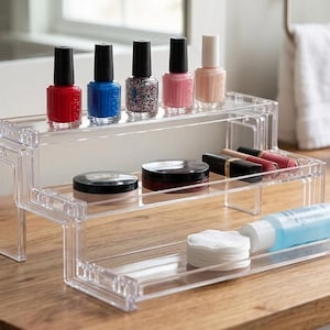 May include: A clear acrylic tiered display stand featuring three levels for organizing cosmetics. The top shelf holds five nail polish bottles in red, blue, glitter, pink, and nude. The middle shelf contains two round compacts and two lipsticks. The bottom shelf holds a stack of white cotton rounds and a small blue bottle.