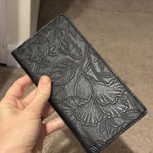 May include: A black leather wallet with an embossed floral design. The wallet is rectangular and features a detailed pattern of leaves and flowers across its surface. The wallet is held in a hand.