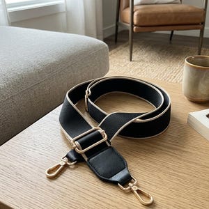 May include: Black and beige bag strap with gold-colored metal hardware. The strap is made of a woven material with a leather accent. The hardware includes two swivel clasps and a buckle.