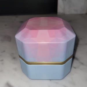 May include: A geometric-shaped box with a faceted top, transitioning from pink to blue. The base is light blue, with a gold band separating it from the top. The box is likely for jewelry or small items.